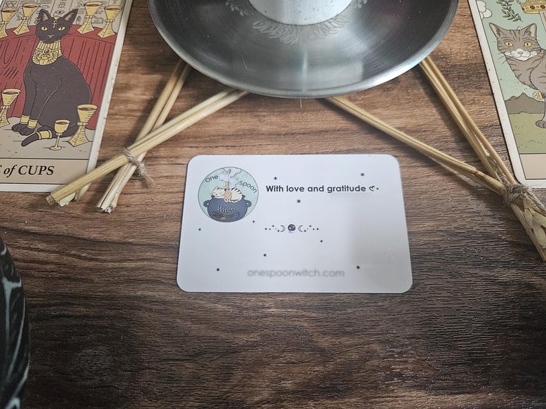 May include: A white rectangular card featuring a circular logo with a cat in a cauldron, the text "One Spoon", "With love and gratitude", and "onespoonwitch.com". The card is decorated with small black stars and moon phase symbols.