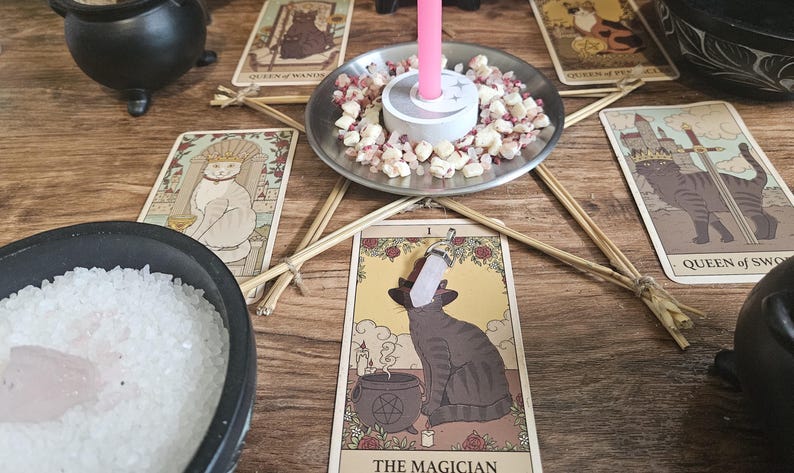 May include: A collection of tarot cards featuring cat illustrations, including The Magician, Queen of Wands, and Queen of Swords. A pink candle sits in a metal dish filled with small crystals, surrounded by a pentagram made of twigs and a bowl of salt.