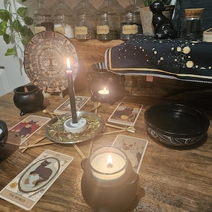 May include: A mystical scene featuring lit candles, tarot cards, and a black cauldron-shaped candle holder. A wooden table displays a circular wooden plaque, jars with labels, and a black cat figurine, creating a witchy aesthetic.