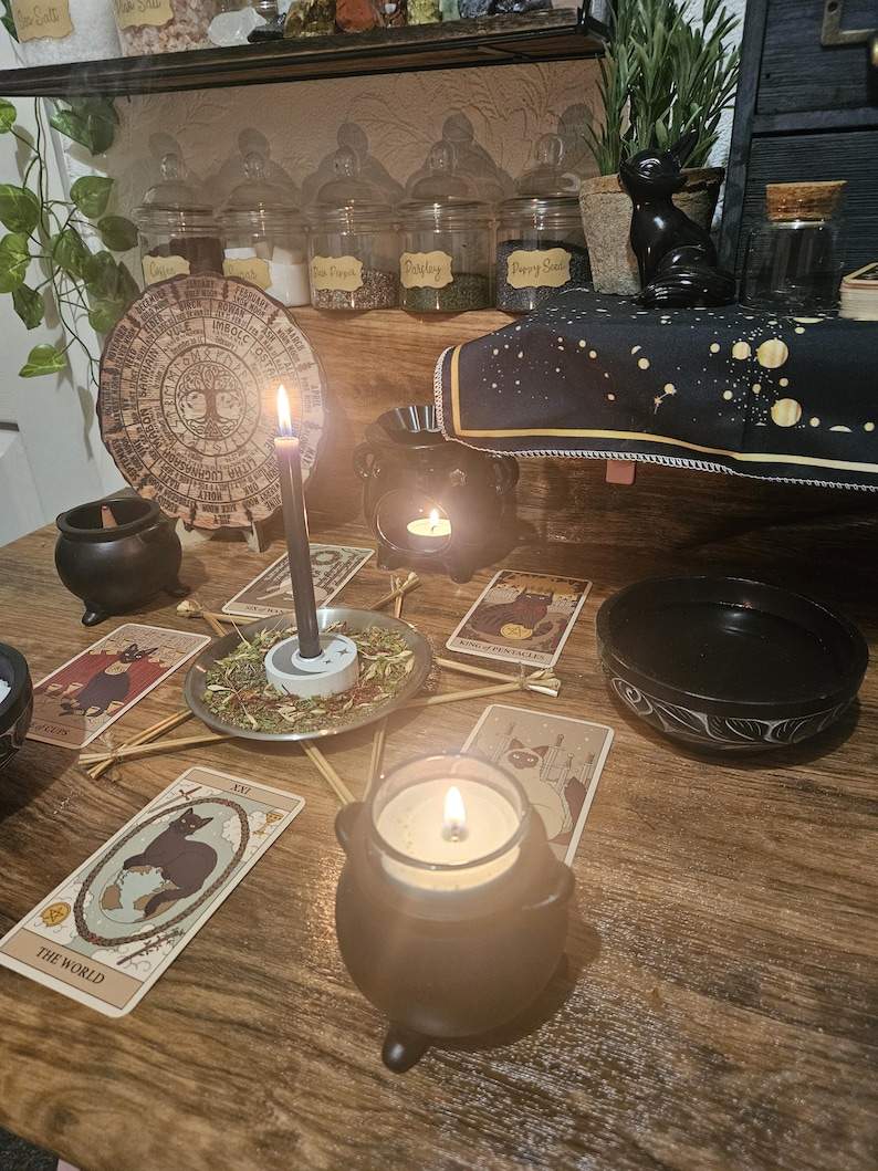 May include: A mystical scene featuring lit candles, tarot cards, and a black cauldron-shaped candle holder. A wooden table displays a circular wooden plaque, jars with labels, and a black cat figurine, creating a witchy aesthetic.