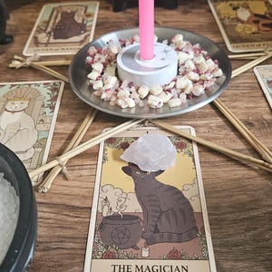 May include: A ritual setup featuring a central silver plate holding a pink candle surrounded by small quartz crystals. Five tarot cards are arranged in a star pattern around the plate, with a black bowl of white salt and a clear quartz point nearby.