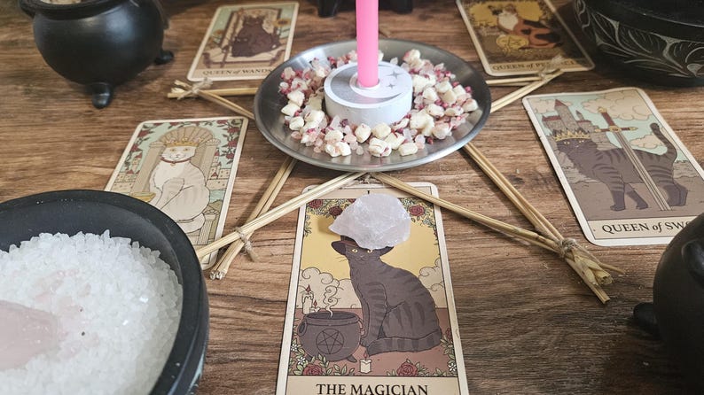 May include: A ritual setup featuring a central silver plate holding a pink candle surrounded by small quartz crystals. Five tarot cards are arranged in a star pattern around the plate, with a black bowl of white salt and a clear quartz point nearby.