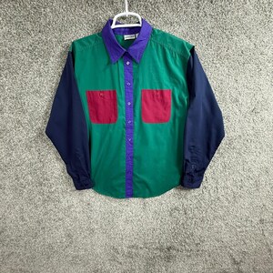 Puede incluir: Una camisa de manga larga con botones que presenta un diseño de bloques de color con cuerpo verde, mangas azul oscuro y cuello y tapeta de botones de color morado. La camisa incluye dos bolsillos en el pecho de color magenta y una percha blanca.