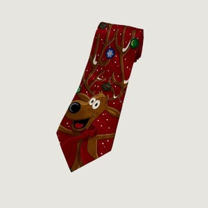 May include: A red necktie featuring a cartoon reindeer with a red scarf, brown antlers decorated with green and blue Christmas ornaments, and a pattern of white snowflakes scattered across the fabric against a solid red background.