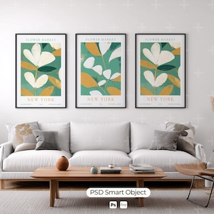 May include: Three framed art prints with floral designs. Each print features a white flower with yellow and green accents against a teal background. The text "FLOWER MARKET NEW YORK" is at the top and bottom of each print. The prints are above a white sofa.