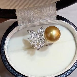 May include: A ring featuring a large, round, golden-colored pearl and a silver-toned floral motif encrusted with numerous small, clear, sparkling stones. The ring is displayed inside a dark blue, oval-shaped jewelry box with a white interior lining.