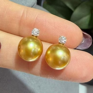 May include: A pair of golden pearl stud earrings. Each earring has a large, round, lustrous golden pearl with a small, sparkling, clear gemstone cluster on top. The earrings are held up against a blurred background.