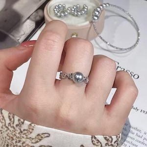 May include: A silver ring with a grey pearl is worn on a finger. A matching bracelet and earrings are displayed in a pink jewellery box. The background includes a patterned fabric and a printed page.