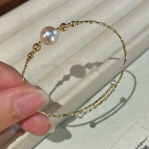 May include: A gold-toned wire bangle bracelet featuring a single large, lustrous, pale pinkish-white pearl accented by small gold beads. The thin, textured metal band wraps around to display two additional tiny white pearls at the open ends.
