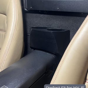 Może przedstawiać: Czarna konsola samochodowa z dwoma uchwytami na kubki, umieszczona między beżowymi skórzanymi fotelami samochodowymi z czerwonymi przeszyciami. Konsola jest przeznaczona do modelu MX-5 NA z lat 1989-1997. Widoczny jest tekst "Perfect Fit for MX-5 NA (1989-1997)".