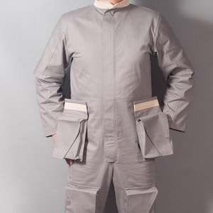 May include: A light gray jumpsuit with multiple pockets and a stand-up collar. The long-sleeved garment has a tailored fit and features rectangular pockets on the chest and legs. The jumpsuit is made of a durable fabric.