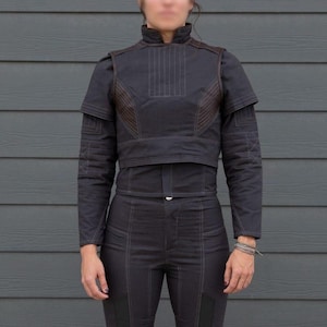 May include: Black two-piece outfit with a high collar and long sleeves. The jacket has brown accents and vertical stitching. The pants are fitted with knee patches and vertical stitching. The outfit is made of a dark fabric.