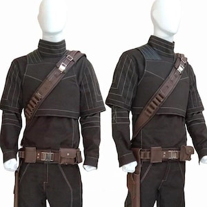 May include: Two black costumes displayed on mannequins. Each costume includes a long-sleeved top with white stitching, matching pants, a brown belt with pouches, and a shoulder strap with ammunition holders. Brown holsters and knee pads complete the look.