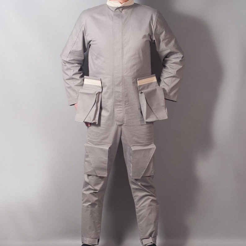 May include: A light gray jumpsuit with multiple pockets and a stand-up collar. The long-sleeved garment has a tailored fit and features rectangular pockets on the chest and legs. The jumpsuit is made of a durable fabric.