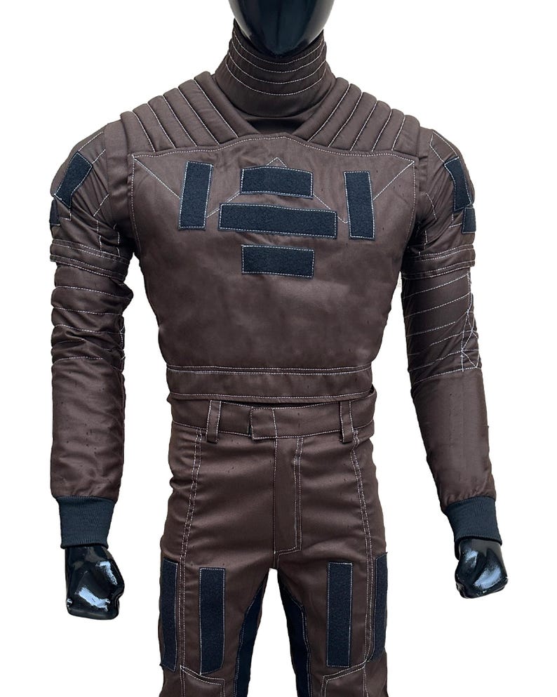 May include: A brown jumpsuit with black accents, displayed on a mannequin. The suit features a high collar, quilted detailing, and multiple black rectangular patches. White stitching outlines the seams and panels. The sleeves end with black cuffs.