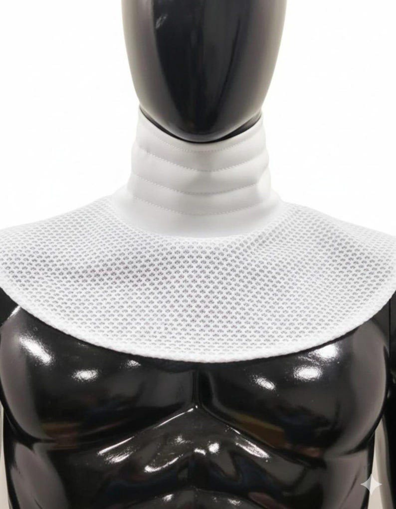 Może przedstawiać: Biały kołnierz szermierczy, z wyściełanym kołnierzem i siatkowym śliniakiem. Kołnierz ma chronić szyję i klatkę piersiową podczas szermierki. Kołnierz jest prezentowany na czarnym manekinie.