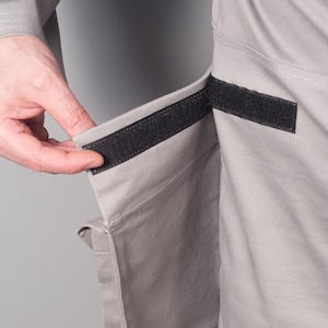 Puede incluir: Primer plano de tela gris claro con un cierre de velcro negro. La tela parece ser parte de una prenda, con una mano que alcanza para asegurar el cierre. La prenda tiene un bolsillo.