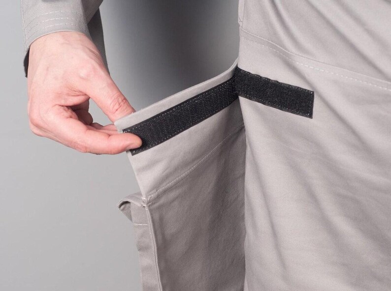 Puede incluir: Primer plano de tela gris claro con un cierre de velcro negro. La tela parece ser parte de una prenda, con una mano que alcanza para asegurar el cierre. La prenda tiene un bolsillo.