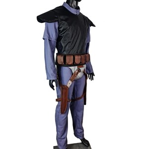 K&ouml;nnte beinhalten: Ein komplettes Kost&uuml;m, das auf einer Schaufensterpuppe ausgestellt ist. Das Kost&uuml;m umfasst einen blauen Overall, eine schwarze Weste mit Schulterpolstern, einen braunen G&uuml;rtel mit Taschen und eine Halterung mit einem Messer. Die Schaufensterpuppe steht auf einer grauen Plattform.
