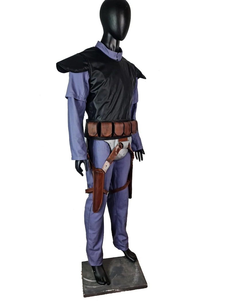 K&ouml;nnte beinhalten: Ein komplettes Kost&uuml;m, das auf einer Schaufensterpuppe ausgestellt ist. Das Kost&uuml;m umfasst einen blauen Overall, eine schwarze Weste mit Schulterpolstern, einen braunen G&uuml;rtel mit Taschen und eine Halterung mit einem Messer. Die Schaufensterpuppe steht auf einer grauen Plattform.