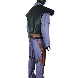 K&ouml;nnte beinhalten: Ein Kost&uuml;m auf einer Schaufensterpuppe, bestehend aus einem hellblauen Overall, einer schwarzen Weste und einem braunen Lederg&uuml;rtel mit Holster. Das Kost&uuml;m umfasst braune Lederbeinriemen und schwarze Handschuhe. Die Schaufensterpuppe steht auf einer grauen Plattform.