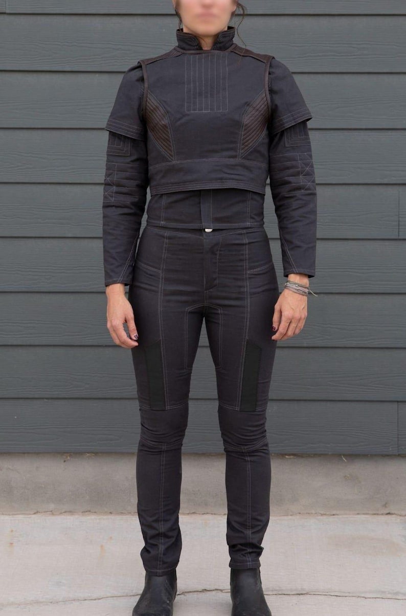 May include: Black two-piece outfit with a high collar and long sleeves. The jacket has brown accents and vertical stitching. The pants are fitted with knee patches and vertical stitching. The outfit is made of a dark fabric.