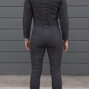 May include: Dark gray two-piece outfit, featuring a jacket with shoulder detailing and a high collar, paired with matching pants. The outfit has visible stitching and a tailored fit. The person is wearing black boots.