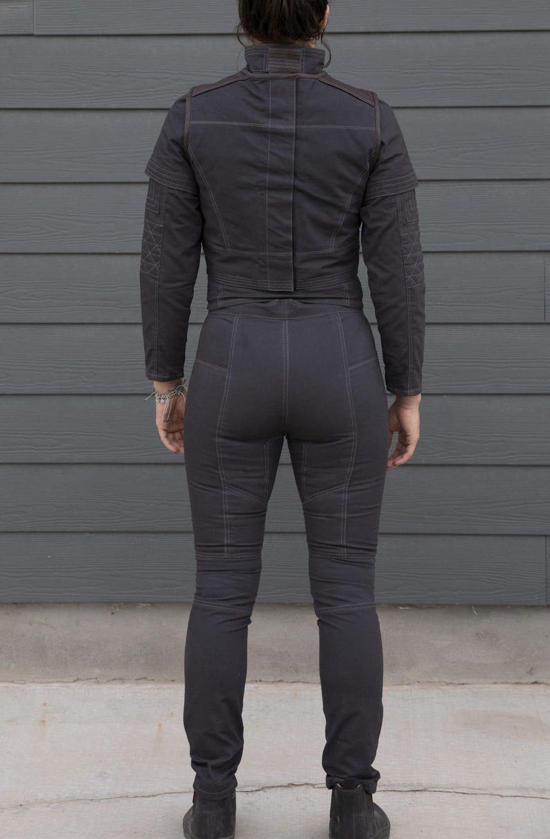 May include: Dark gray two-piece outfit, featuring a jacket with shoulder detailing and a high collar, paired with matching pants. The outfit has visible stitching and a tailored fit. The person is wearing black boots.