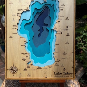 May include: A wooden Lake Tahoe bathymetric map featuring layered blue cutouts representing water depth. The light wood frame displays geographic labels, a compass rose, and coordinates. Text includes Latitude 38.939926, Longitude -119.977187, and Lake Tahoe California & Nevada, USA.