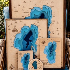 May include: A collection of wooden Lake Tahoe bathymetric maps in various sizes, featuring layered blue water depths, engraved geographic details, coordinates, and compass roses. A circular wooden ornament with a heart icon is included.