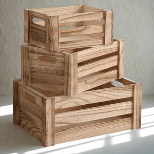 Op de afbeelding: Drie houten kisten van verschillende formaten zijn op elkaar gestapeld. De kisten zijn gemaakt van lichtbruin hout met zichtbare nerf en hebben uitgesneden handgrepen. Het ontwerp omvat horizontale lamellen voor ventilatie, wat een rustieke uitstraling creëert.