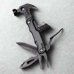 Könnte beinhalten: Ein Multifunktionswerkzeug mit Hammer, Zange, Messer und Schraubendreher-Bits. Das Werkzeug besteht aus Edelstahl und hat eine grau-schwarze Oberfläche. Der Hammerkopf befindet sich oben, Messer und Zange sind ausgeklappt. Auf hellem, grauem Hintergrund.