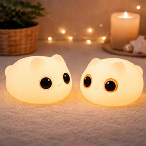 May include: Two glowing cat-shaped night lights with large black eyes and orange accents. The lights are a soft white color and are sitting on a textured surface. A lit candle and decorative lights are in the background.