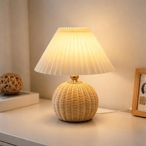 May include: A decorative table lamp with a woven rattan base and a pleated cream-colored lampshade. The lamp has a warm, inviting glow, perfect for a bedroom or living room. The lamp is on a white surface.