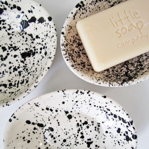 May include: Three white ceramic soap dishes with black splatter paint. The middle dish has a bar of soap inside that says "little soap company".