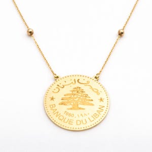 May include: A gold-colored necklace featuring a round pendant with a detailed tree design and Arabic text. The pendant is inscribed with "BANQUE DU LIBAN" and the year "1960". The necklace has a delicate chain with small gold beads.