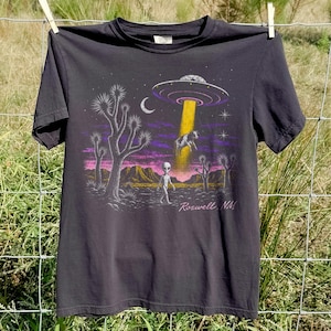 Könnte beinhalten: Ein anthrazitfarbenes Kurzarm-T-Shirt mit einem Grafikdruck einer Wüstenlandschaft, einem UFO, das eine Kuh entführt, einer Alien-Figur, einer Mondsichel und dem Schriftzug Roswell, NM in Schreibschrift vor einem violetten und rosafarbenen Himmel.