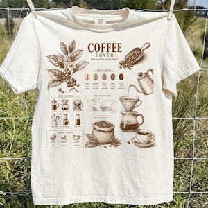 May include: A cream-colored short-sleeve t-shirt featuring a brown graphic print titled COFFEE LOVER. The design includes illustrations of a coffee plant, roast levels, brewing methods, a world map, a coffee bag, and various coffee brewing equipment.