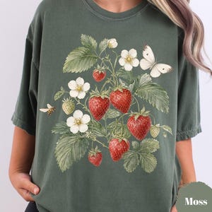 May include: A moss green short-sleeve t-shirt featuring a detailed botanical illustration of red strawberries, white strawberry blossoms, green leaves, a small bee, and a white butterfly centered on the front of the garment.