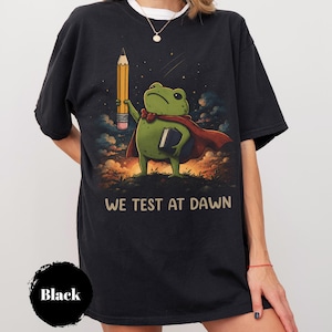 May include: A black t-shirt featuring a graphic of a green frog wearing a red cape, holding a yellow pencil and a book. The background shows a sunset sky with clouds. The text "WE TEST AT DAWN" is printed in beige below the frog illustration.