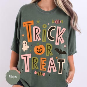 Może przedstawiać: Mchowo-zielony t-shirt z krótkim rękawem z napisem TRICK OR TREAT wykonanym dużymi, kolorowymi literami. Wzór zawiera pomarańczową dynię, białego duszka, czarnego nietoperza oraz dwa cukierki, ozdobione małymi gwiazdkami.