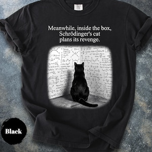 Può includere: Una t-shirt nera a maniche corte con la grafica di un gatto nero seduto in un angolo di fronte a una parete coperta da complesse equazioni e diagrammi di fisica. Il testo sopra la grafica recita: "Meanwhile, inside the box, Schrödinger's cat plans its revenge."