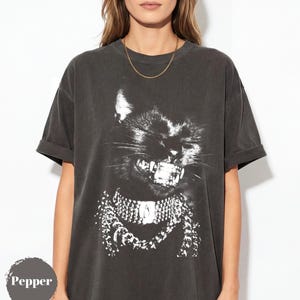 May include: Dark gray t-shirt featuring a black cat graphic with a detailed, open-mouthed expression and a decorative collar. The cat graphic is in white, contrasting against the shirt's color. The shirt has short sleeves and a relaxed fit.