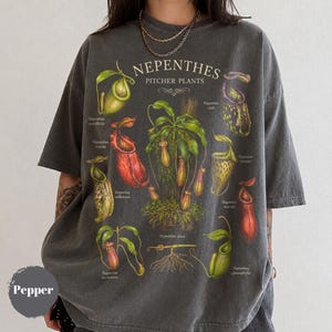 以下が含まれることがあります： チャコールグレーの半袖Tシャツ。ネペンテス（ウツボカズラ）の様々な植物のボタニカルイラストが描かれています。グラフィックには「NEPENTHES PITCHER PLANTS」という文字と、ヴィンテージ風のデザインで各植物種のラベルが記されています。
