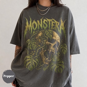 May include: A charcoal gray short-sleeve t-shirt featuring a graphic print of a skull intertwined with green monstera leaves. The text MONSTERA is printed in a stylized green font above the skull. A circular gray icon with the text Pepper is visible.