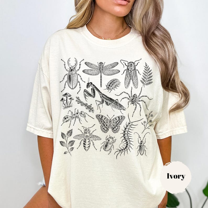 May include: An ivory short-sleeve t-shirt featuring a detailed black ink illustration of various insects, including a praying mantis, dragonfly, beetle, butterfly, spider, and centipede, arranged in a collage pattern across the front chest area.