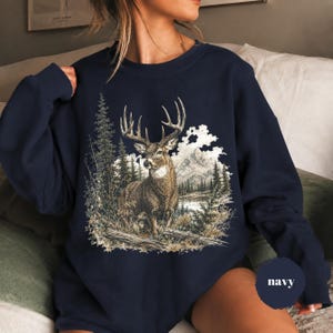 K&ouml;nnte beinhalten: Ein marineblaues Sweatshirt mit Rundhalsausschnitt und einem detaillierten Grafikdruck eines braunen Hirsches mit gro&szlig;em Geweih in einer Waldlandschaft mit hohen Kiefern, einem ruhigen See und fernen Bergen unter bew&ouml;lktem Himmel, mit dem Wort navy.