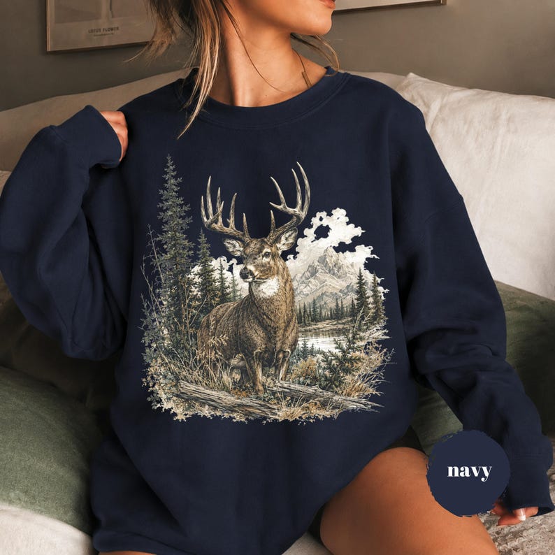 K&ouml;nnte beinhalten: Ein marineblaues Sweatshirt mit Rundhalsausschnitt und einem detaillierten Grafikdruck eines braunen Hirsches mit gro&szlig;em Geweih in einer Waldlandschaft mit hohen Kiefern, einem ruhigen See und fernen Bergen unter bew&ouml;lktem Himmel, mit dem Wort navy.