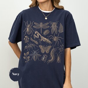 May include: A navy blue t-shirt featuring a detailed gold-toned graphic print of various insects, including a beetle, dragonfly, praying mantis, spider, butterfly, and centipede, along with small botanical elements and mushrooms.