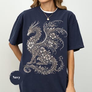 May include: A navy blue short-sleeve t-shirt featuring a large, intricate graphic of a dragon composed entirely of detailed floral patterns and leaves in shades of cream and light gray. The design covers the center of the chest area.